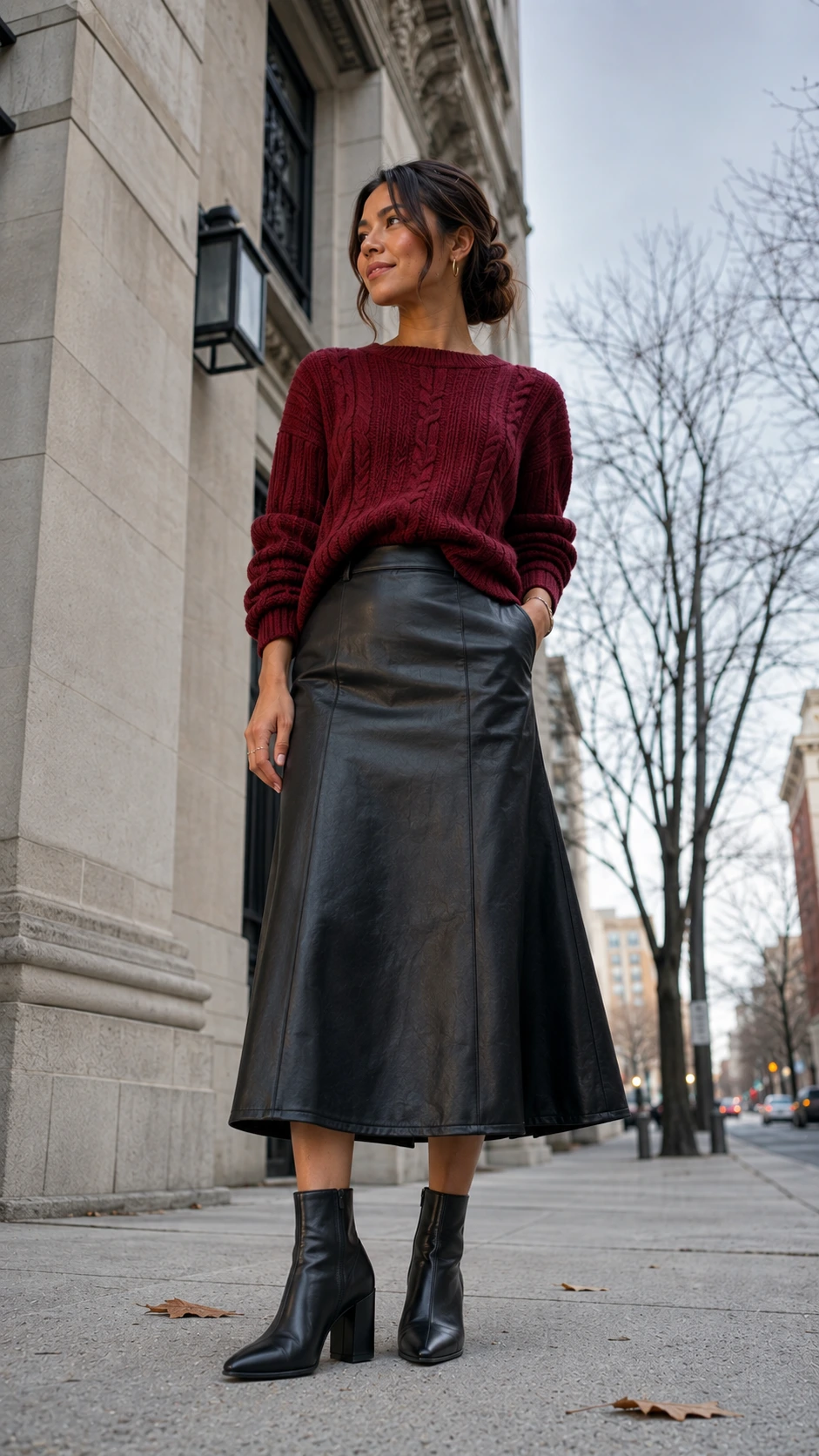 Woman wearing a chunky cranberry red ribbed knit sweater tucked into a sleek black leather midi skirt, styled for a festive work dinner outfit on a city sidewalk